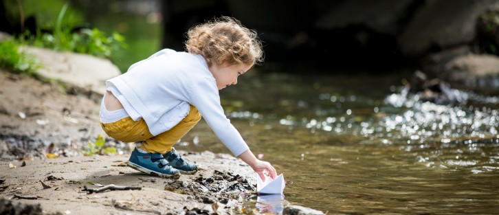 A toddler by a stream with a paper boat.