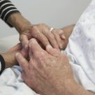 An elderly couple are holding hands in a hospital bed.